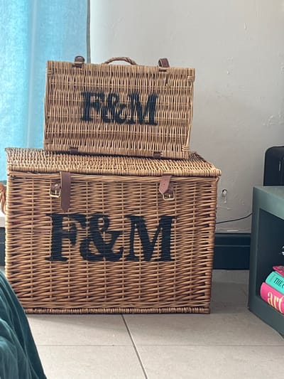 The image shows two brown wicker baskets with black lettering stacked on top of each other. The letters on the baskets appear to spell out "F&M". The baskets have leather straps with buckle closures. The baskets are set against a backdrop of a light blue and white window in the lower left and a light gray wall in the background. Part of a green item, possibly clothing or upholstery, is visible in the lower right corner. A dark gray shelf with books on top is positioned above the baskets. The location in Liverpool, United Kingdom is not identifiable.