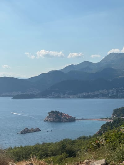 The image depicts a scenic coastal view from a hillside overlooking the Adriatic Sea in Blizikuće, Montenegro. In the foreground, lush green trees and dry brush cover the slope. The middle ground is dominated by the clear blue sea, where several boats are visible, including two small motorboats and a few other vessels. A notable feature is the small island of Sveti Stefan, connected to the mainland by a narrow causeway, with its cluster of red-roofed buildings. Adjacent to Sveti Stefan, a sandy beach is visible. Further in the background, a town with numerous buildings, including some larger modern structures, sprawls along the coastline. Behind the town, a series of majestic, verdant mountains rise, their peaks softened by distance and a light haze. The sky is a serene pale blue with scattered, wispy white clouds. The lighting suggests a bright, sunny day, casting a gentle glow over the landscape.