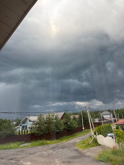 This media file captures a rural residential scene in Kiskelovo, Russia, under dramatic, stormy skies. The image is oriented vertically, with the dramatic clouds dominating the left half and a village road lined with houses and vegetation on the right.
The sky is filled with dark, heavy storm clouds, broken by shafts of sunlight that create a sense of an impending downpour or a brief respite from it. The weather appears turbulent, with a moody atmosphere.
On the right side of the image, a gravel road curves through a residential area. Several houses are visible, characterized by their pitched roofs and wooden fences. One prominent house is painted light blue with white trim and features several windows and a porch. Trees and lush green grass surround the houses, indicating a summer setting. Power lines crisscross the scene, supported by utility poles. A fence with a heart-shaped decorative element is visible near the road.
The overall scene evokes a sense of quiet rural life under the influence of dramatic weather, possibly late afternoon or early evening given the lighting conditions and cloud formations. There is no visible text in the image.