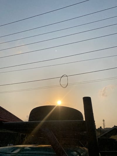 The image presents a view looking up towards the sky, partially obscured by a dark, metallic structure on the right. Thin, parallel electrical wires stretch across the frame, converging towards the left. A circular, possibly hanging object is visible on one of the wires. The sun, appearing as a bright, small circle, shines through the sky, creating a slight flare effect. The sky's gradient transitions from light grey-blue on the left to a warm, hazy orange-yellow on the right, around the sun.