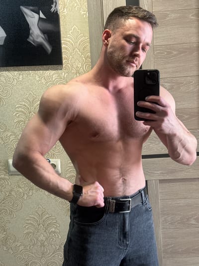 A man takes a selfie indoors. He is shirtless, wearing jeans with a black belt and a black wristwatch. The man is holding a black smartphone. The background includes a door with a wood-grain finish and a wall with patterned wallpaper. A black and white artwork is partially visible on the wall. The specific location is indoors, and it is not possible to definitively confirm if it's Dobryanka, Russia from the image alone.