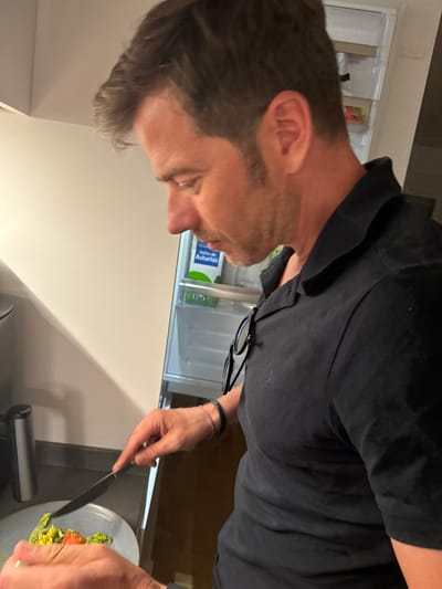 A man is shown from the chest up, wearing a black polo shirt. He is standing in what appears to be a kitchen, with a refrigerator visible in the background. He is in the process of preparing food, as he holds a knife over a plate containing lettuce and what looks like a tomato. His right hand is visible, holding the knife, and his left hand is near the plate. He has short brown hair and a light beard. The scene is brightly lit, suggesting it is daytime or well-lit indoors. The man's focused expression indicates he is concentrating on his task.