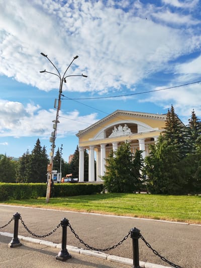 The image depicts the National Art Gallery in Yoshkar-Ola, Russia, on a bright, cloudy day. The building features a neoclassical facade with tall white columns and a pediment adorned with sculptures. In front of the gallery, a lush green lawn is bordered by a neatly trimmed hedge. A paved walkway runs parallel to the lawn, separated by a black chain fence with ornate posts.

A tall, multi-branched street lamp stands prominently in the foreground, equipped with multiple lights and various antennas or communication devices. Behind the hedge and to the left of the building, a few evergreen trees are visible, along with some taller buildings in the distance. To the right of the building, more evergreen trees create a dense border. A bright blue sky with fluffy white clouds fills the upper portion of the image.

There are no people visible in the scene. The focus is on the architecture, the surrounding landscape, and the prominent street lamp. The presence of the street lamp with communication equipment suggests it might be a functional fixture rather than purely decorative. The scene appears serene and well-maintained, characteristic of a public space in a city. The time of day appears to be daytime, given the bright natural light.