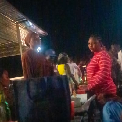 The image, likely taken at night in or near Ube, Nigeria, shows a gathering of people under a corrugated metal roof. Two women are prominent in the foreground: one in a brown top and head wrap, the other in a red and white striped top. Other individuals are visible in the background, some wearing yellow or white tops. There are blurred objects in the foreground, possibly tables or containers, and a green bottle is visible on the left. The lighting is dim, suggesting an outdoor setting at night. The overall impression is of a casual social event or gathering.