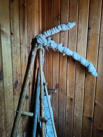 A close-up shot captures a collection of wooden tools leaning against a rustic wooden wall. The wall is constructed from vertically oriented planks, exhibiting a natural wood grain with variations in color from light brown to a richer, darker tone. Knots are visible in the wood, adding to its textured appearance.
Dominating the center of the frame is a large, curved object that appears to be a tool or a part of one. It is composed of two wooden poles joined at an angle, with the longer section extending upwards and to the right. This prominent piece is wrapped in a white, somewhat tattered fabric that is twisted around it, creating a textured, rope-like effect. The fabric has frayed edges and is loosely secured, suggesting it might be a protective covering or a decorative element.
To the left and below this main piece, other wooden objects are visible. There's a tripod-like wooden structure with two legs visible, made of rough-hewn wood. A pointed wooden stake, darker in color and with a weathered appearance, leans against the main tool. Another long, thin wooden pole is also present. Some wooden dowels or pegs are attached to the wooden wall itself, serving as hooks. A glimpse of a blue quilted fabric or material is visible behind the wooden objects on the lower right.
The lighting in the scene is somewhat dim, casting subtle shadows on the wall and highlighting the textures of the wood and fabric. The overall impression is one of rustic craftsmanship or perhaps a display of traditional tools in a rural or historical setting.
This scene is located in the city of Semenkovo, Russia, likely within a museum or historical exhibit dedicated to rural life and traditions. The wooden wall and the style of the tools suggest an authentic representation of past agricultural or crafting practices in the region.