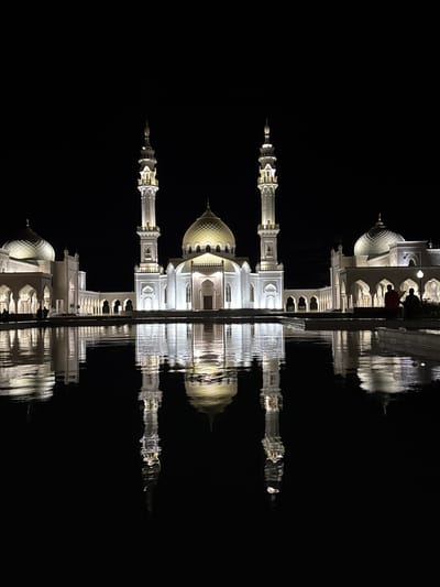 A nighttime exterior shot captures the White Mosque in Bolgar, Russia, illuminated against a dark sky. The mosque, a grand white structure, features multiple domes and two tall minarets. Its architecture is intricate, with arched windows and decorative elements, all brightly lit. In front of the mosque is a large, still body of water that perfectly reflects the illuminated building, creating a symmetrical and striking visual effect. The water is dark, emphasizing the reflections of the mosque and the lights. On the right side of the image, two figures are visible near the water's edge, silhouetted against the mosque's light. One person is wearing a red shirt, and the other is seated, their forms indistinct in the low light. The scene conveys a sense of grandeur and tranquility.