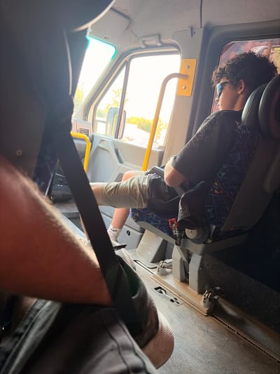 The image shows the interior of a vehicle, likely a bus or van, from a first-person perspective. A young person with curly dark hair and wearing sunglasses is seated in a seat, facing away from the camera and looking out a window. They are wearing a dark t-shirt with a patterned design and light-colored shorts. The seat itself has a dark leather headrest and a seat cushion with a blue and black abstract pattern.

The vehicle's interior is utilitarian, with grey plastic panels and a visible yellow grab handle. Sunlight streams in through the window, suggesting it is daytime. The outside view is blurred but shows greenery and trees, indicating an outdoor setting.

In the foreground, a black seatbelt is prominently displayed, and part of another person's arm and leg, also wearing a seatbelt, are visible, suggesting the camera holder is also a passenger. The overall mood appears relaxed, with the seated person seemingly engaged with the view outside. No visible text is present in the image. The location context is Příbram, Czechia.