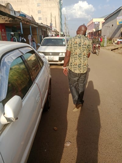 A man wearing a patterned shirt and blue jeans walks away from the camera down a paved street in Kapsabet, Kenya. He is in the foreground of the image, and his long shadow is cast in front of him. A white sedan is parked on the right side of the road, with a red stripe running down the side. The car has a license plate that reads "KCL 802U." In the background, there are other buildings, people, and a motorcycle. The scene appears to be during the daytime and the weather is sunny.
