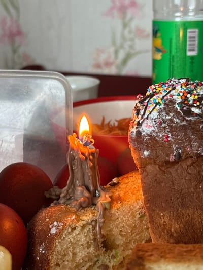 A close-up, high-angle shot depicts a section of Easter bread, likely a Kulich, adorned with colorful sprinkles on top. The bread is partially cut, revealing its interior. A lit candle with elaborate, melted wax flows down the side of the bread. Several red-brown dyed Easter eggs are situated beneath the bread. The setting appears to be indoors, possibly a kitchen or dining area. A bottle, a floral-patterned surface, and a portion of green packaging are visible in the background. The specific location is Пожарское сельское поселение, Ukraine.