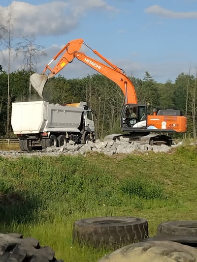 An orange Hitachi excavator is dumping rocks into the bed of a white and silver dump truck. The excavator's arm is extended over the truck, with its bucket tilted, releasing a stream of rocks. The dump truck is a large, multi-wheeled vehicle, positioned to receive the load. Both vehicles are situated on a rough, rocky surface, with a grassy embankment in the foreground. In the background, a dense forest of trees is visible under a bright, partly cloudy sky. The image appears to be taken in an outdoor construction or excavation site, potentially in a rural or semi-rural area of Novoselytsia, Ukraine, given the context. There is no discernible human activity beyond what might be inside the excavator's cabin.