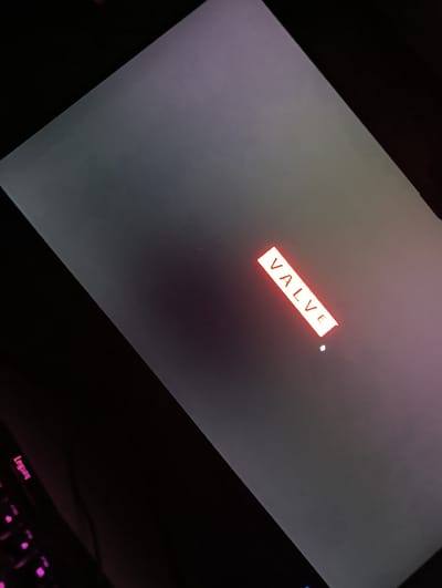 The image shows a laptop screen displaying the "Valve" logo, a common splash screen for Valve Corporation's software, including Steam. The logo is presented in a rectangular red and white box, with the word "VALVE" in white text on a red background. The screen itself appears to be displaying a light gray background with a gradient effect, possibly due to ambient lighting or screen settings. The laptop is likely in a dark room, as suggested by the illuminated keyboard in the bottom left corner, which has pink backlighting and displays the "Legion" logo. The overall scene is dark, with the exception of the laptop screen and keyboard. The image is taken from an angle, and the screen is slightly tilted. There are no people visible in the image. The time of day and specific location within Dushanbe, Tajikistan, cannot be determined from the visual information provided. The activity taking place is the booting up or launching of a program on the laptop, indicated by the Valve logo.