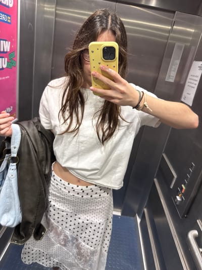 A young woman with long brown hair is taking a selfie in an elevator. She is holding a yellow iPhone and wearing a white t-shirt, a polka dot skirt, and a jacket. She also has bracelets on her wrist. The elevator has stainless steel walls and a blue checkered floor. There is a display on the wall with multiple controls. A pink box of board games can be seen in the reflection. While the precise location in Hull is not directly evident from the image, the presence of an elevator suggests an indoor, possibly public, setting.