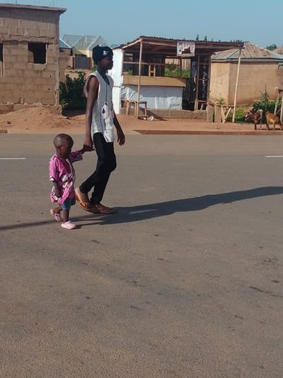 In Tumfure, Nigeria, a young child in a pink patterned shirt and denim shorts, wearing pink Crocs, walks hand-in-hand with a taller individual wearing a white sleeveless top and black pants, and a black cap. They are walking on a paved road. In the background, there are buildings under construction, a stall with a tin roof, and two goats. The scene suggests a typical street environment in a Nigerian town, with a blend of residential and commercial structures. The lighting and shadows indicate it is daytime.