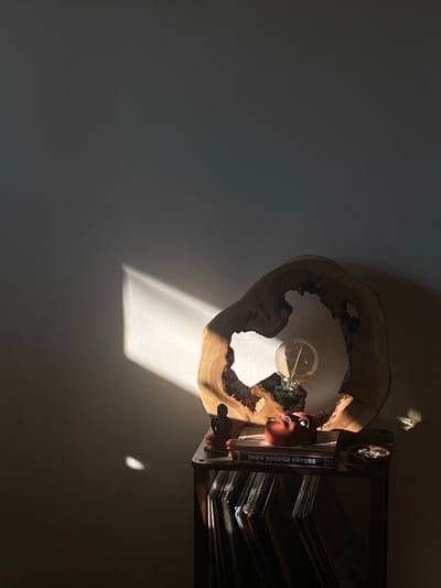 The image shows a dimly lit room, possibly a home or studio, in Shebunino, Russia. The scene is dominated by strong natural light filtering in from the left, casting a sharp diagonal beam across the wall and onto a wooden surface. This wooden surface appears to be part of a unique lamp, made from a slice of a tree trunk with a natural, irregular opening. Inside this opening, a vintage-style lightbulb is visible, emitting a warm glow.
To the right of the lamp, a black shelf unit is partially visible, filled with what appear to be vinyl records. One stack of records has a visible spine with text that reads "1000 RECORD COVERS." A small, reddish-brown, cartoonish figurine is placed on the shelf, appearing to interact with the lamp. The overall mood is serene and contemplative, with the play of light and shadow creating a dramatic effect. The time of day is suggested to be late afternoon or early morning, given the long shadows and the angle of the sunlight. No specific weather conditions are apparent, but the strong light implies a clear day.