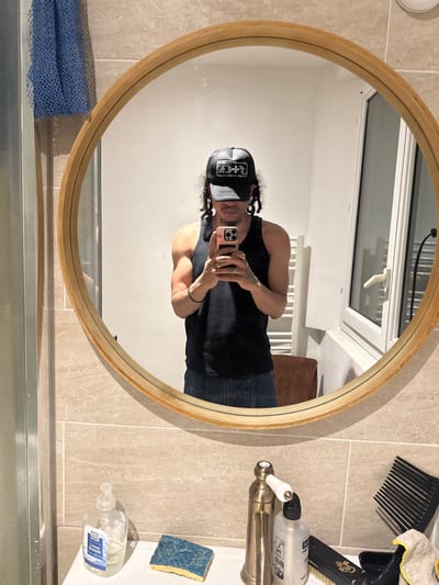 A person wearing a black tank top and a black baseball cap is taking a selfie in a circular mirror. The bathroom setting includes beige tile walls, a window with white framing, a sink with a brass faucet, and various toiletries and cleaning supplies. There is no evidence to suggest this image was taken in Massy, France.