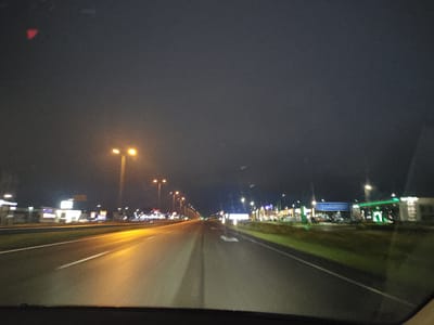 The image shows a nighttime view of a highway in Laagri alevik, Estonia. The road is wet, reflecting the orange glow of streetlights. On the right side of the road, there are several commercial buildings, including a brightly lit gas station with green accents. On the left side, more illuminated buildings are visible, suggesting a commercial area. The sky is dark and overcast. The perspective is from inside a vehicle, looking forward. There are no people or animals clearly visible in the frame, and no discernible actions or interactions are taking place. The overall impression is one of driving through a well-lit, developed area at night.