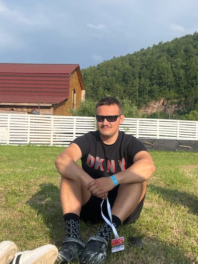 A young man is sitting on the grass in what appears to be a park or an outdoor event. He is wearing a black t-shirt with red lettering and black shorts. He has on black and white patterned socks and black Crocs. A blue wristband is on his left wrist, and a lanyard with a badge is hanging from his right hand. He is wearing sunglasses.
In the background, there is a wooden building with a red roof and a white wooden fence. Behind the fence, there is a steep, forested hill. The sky is clear and blue, suggesting it is daytime with good weather. The grass is green and appears well-maintained. The man's posture is relaxed, and he seems to be enjoying himself.
The text "CONCE" is visible on the badge hanging from the lanyard. The location context provided is Solontsy, Russia. This suggests the image was taken in Solontsy, Russia, possibly during an outdoor festival or gathering.