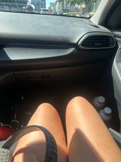 The image shows the interior of a car, likely in Zurich, Switzerland, during the daytime. The sun is shining brightly, casting shadows across the dashboard and the legs of a person sitting in the passenger seat. The person's legs are bare and appear to be tanned, suggesting warm weather. They are wearing a dark-colored skirt or shorts.

On the dashboard, a silver-trimmed air vent is visible. Below it, an "AIRBAG" warning label is printed on the plastic. To the right of the air vent, on the passenger side of the car, two white plastic bottles with red and white labels are placed. The labels are too blurry to read any text clearly.

Through the car window on the left, a view of a city street is visible. Buildings and trees can be seen, along with what appears to be traffic. The sunlight suggests it is either morning or afternoon. The overall mood of the image is casual and relaxed, hinting at a journey or a stop during a trip.