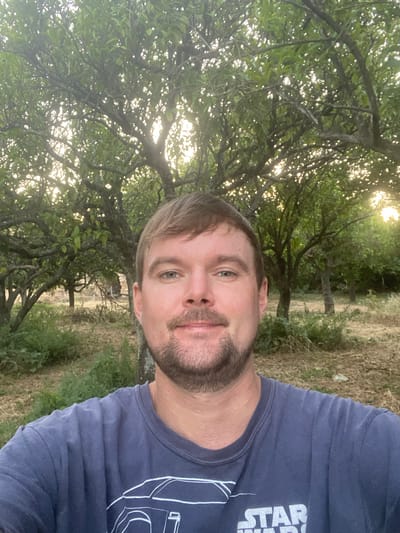 A man with fair skin, brown hair, and a short beard takes a selfie outdoors. He is wearing a dark blue t-shirt with a white outline of a car and the words "STAR WARS" printed on it. He is positioned in the center of the frame, with the lower part of his face, neck, and shoulders visible. Behind him, there are several trees with green foliage, some of which are illuminated by the setting sun, creating a warm, dappled light. The ground is covered with dry grass and fallen leaves. The scene appears to be a garden or orchard. This photo was taken in the city of Olhão, Portugal, specifically within the Parque Natural da Ria Formosa, known for its rich biodiversity and scenic landscapes.