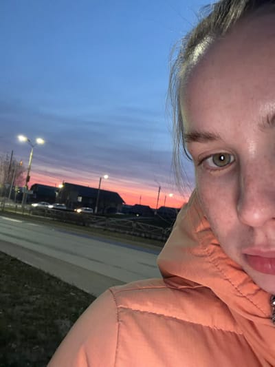 The image is a tilted, close-up selfie or first-person perspective shot taken outdoors at dusk, likely in Berezniki, Russia.

The main subject is a person, likely female, whose face occupies the upper right portion of the frame. Visible features include part of their forehead, a prominent green or hazel eye, part of their nose, and pink lips. Their fair skin shows some minor freckling. Light brown or blonde hair is visible along the left side of their face. The person is wearing an orange or peach-colored puffer jacket, which covers most of their body in the frame, showing texture and stitching.

The background, visible in the lower left portion of the image, features a striking sunset or twilight sky. The sky transitions from a deep blue at the top to vibrant shades of orange and red along the horizon. Silhouettes of buildings, streetlights, and what appear to be distant vehicles are visible against the colorful sky, indicating an urban or suburban setting. A road or pavement and a patch of grass or dry ground can be seen in the lower right corner, adjacent to the person's jacket. The streetlights are illuminated, suggesting dim natural light.

The activity appears to be the person taking a self-portrait with the scenic twilight as a backdrop. There is no visible text in the image.
