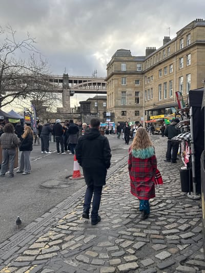 An outdoor scene shows people walking on a cobblestone street in front of buildings. A person wearing a red plaid coat with a teal furry collar walks away from the camera. Another person in a black coat walks in the same direction. Street vendors with awnings and displays are visible. A bridge is partially visible in the background. The sky is overcast.