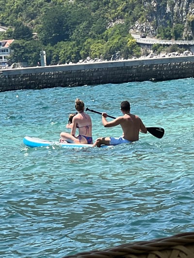 Two individuals are paddleboarding on clear turquoise water. A woman wearing a bikini is seated on the front of the paddleboard, with her legs crossed. Next to her, a younger individual, possibly a child, is seated. A man, shirtless and wearing light-colored shorts, is seated at the rear of the paddleboard and is holding a paddle. The paddleboard is white with blue accents. In the background, a stone wall lines the shore, beyond which are lush green trees and a hillside. A road is visible winding along the hillside, and a few buildings are nestled amongst the trees. The scene appears to be in Risan, Montenegro, characterized by the hilly terrain and the clear Adriatic Sea.