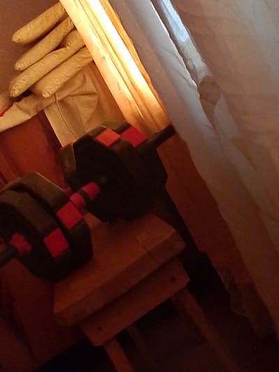 In an indoor setting, possibly a room in Pavlovo, Russia, a pair of dumbbells rests on a wooden bench. The dumbbells have black weight plates with red accents and are positioned on a rustic wooden bench. To the left of the dumbbells, what appears to be a stack of rolled-up items, possibly mats or towels, in a beige color are visible. Behind these items, a warm yellow light source emanates, casting a glow on the scene. A sheer, light-colored curtain hangs on the right side of the frame, partially obscuring the background and contributing to the dimly lit, cozy atmosphere. The overall impression is one of a home gym or a storage area for exercise equipment.