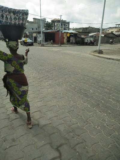 The image depicts an urban street scene in Cotonou, Benin, under an overcast sky. In the foreground, a woman is walking away from the viewer on a road paved with gray interlocking concrete blocks. She wears a long dress featuring green, purple, and yellow spiral patterns, with a dark brown fabric wrapped around her waist, and pink open-toed sandals. A dark-colored basin, covered with a fabric printed with various human hairstyles, is balanced on her head. Her right arm is raised and slightly bent at the elbow, hand open.

The road continues into the middle ground, where it appears to intersect or curve. To the left, a dark-colored sedan is visible, followed by a yellow motorbike and its rider. Behind these vehicles, multiple multi-story buildings with light-colored walls and a taller structure with a brown roof are present. To the right, a series of single-story commercial structures includes a reddish-brown building with yellow clothing displayed externally, and an adjacent smaller, rustic-looking building. Several individuals and parked motorbikes are visible near these structures. Tall, slender utility poles punctuate the scene, and overhead wires are faintly visible against the sky.
