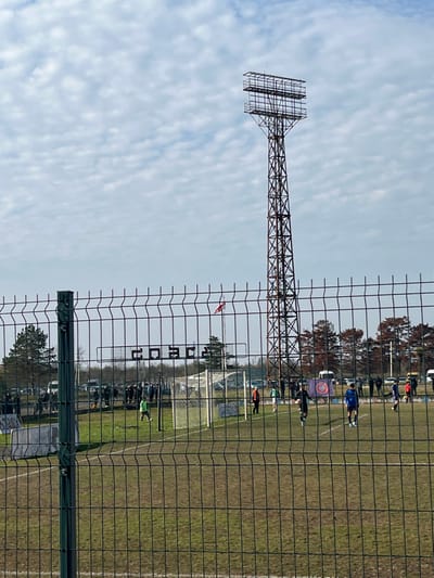 The image depicts an outdoor football (soccer) field located in ქალაქი ლანჩხუთი, Georgia, observed from behind a dark green wire mesh fence. The scene unfolds under a partly cloudy sky.
In the foreground, a prominent dark green wire mesh fence extends across the entire frame, with a vertical fence post visible on the left. Beyond this fence, a brown and green grass pitch is visible. On the left side of the pitch, near a white goal frame, a player in a lime green kit stands near a football. Further into the field, players in black and blue kits are present. Other individuals are distributed around the perimeter of the playing area. A white sign with the word "COACH" in dark lettering is affixed to a structure behind the left goal. A flag displaying a red cross on a white background, consistent with the flag of Georgia, flies from a flagpole to the right of the "COACH" sign. A purple, black, and white banner featuring a circular logo is positioned on the far side of the field.
A tall, rusty-brown metal lattice floodlight tower, characterized by a multi-tiered platform structure at its summit, dominates the right side of the background. Behind the field, parked vehicles, a line of deciduous trees with reddish-brown foliage, and a cluster of green coniferous trees on the far left are discernible. The sky above is light blue with scattered white stratocumulus clouds.