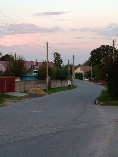 The image depicts a rural road in Смілавіцкі сельскі Савет, Belarus, during the twilight hours. The road is asphalt and curves to the right, flanked by vegetation and residential structures. On the left, a red metal gate is visible, leading to a property with a wooden fence. Further down the road, several houses with reddish-brown roofs and some with green accents are nestled amongst trees. Power lines are strung along poles that line the road. A white car is parked on the side of the road near a yellow house. The sky is a soft gradient of pink and pale blue, indicating the end of daylight. The overall impression is one of quiet, rural life at dusk.