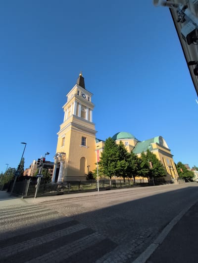 The image depicts the Oulu Cathedral in Oulu, Finland, on a clear, sunny day. The cathedral is a prominent yellow building with a tall, square bell tower topped with a black spire and a golden cross. The tower features a clock face. To the right of the tower, the cathedral has a green domed roof and a long, yellow body. Several lush green trees are planted in front of the building.
In the foreground, a cobblestone street is visible, with a pedestrian crossing marked by lighter-colored stones. Traffic lights are present on the left side of the street, with one light showing red. A black metal fence runs along the perimeter of the cathedral grounds. In the background, other buildings, including an apartment building, can be seen. The sky is a bright, clear blue. There are no people visible in the image. The scene is static and captures the exterior architecture of the cathedral and its surrounding urban environment.