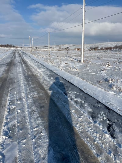 The scene, set in Qoraobdol QFY, Uzbekistan, depicts a snow-covered rural landscape under a partly cloudy blue sky. A two-lane asphalt road, partially cleared but still covered with snow, ice, and slush, stretches from the foreground into the distance, exhibiting prominent vehicle tire tracks. In the immediate foreground, a long, dark, human-shaped shadow is cast across the road from the bottom center-right. Both sides of the road are covered in a uniform layer of white snow, interspersed with sparse, low-lying, dormant vegetation. Along the right side of the road, a series of light-colored wooden utility poles with overhead power lines extends into the background. The midground and background feature gently rolling hills, also dusted with snow. Scattered small, light-colored buildings and bare deciduous trees are visible on these distant slopes. The overall lighting indicates strong sunlight, creating distinct shadows across the snow.