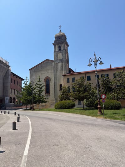 The image shows a street scene in Pisa, Italy, on a clear day. A grey asphalt road curves to the right in the foreground, bordered by bollards. On the left side of the road, a building with scaffolding is visible. In the center and right of the image, a church with a bell tower stands next to a building with multiple windows. Trees and green grass are present in front of the buildings. A street lamp with decorative scrollwork is located on the right side of the road, with a "30" speed limit sign and a "no parking" sign below it. The sky is a clear blue.