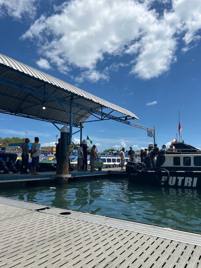 In the city of Tidore Islands, Indonesia, a busy pier is depicted on a clear, sunny day. The foreground shows a metallic walkway with a textured surface, leading towards the water. Several people are gathered on the pier, some standing under a covered shelter with a corrugated metal roof supported by blue metal beams and pillars. Others are by the water's edge, near several boats. One boat, labeled "PUTRI," is prominent, with a white Indonesian flag flying from its mast. People are seen interacting with the boat, possibly embarking or disembarking. The water is a deep blue-green, reflecting the sky and clouds. In the background, more boats and buildings are visible, suggesting a bustling port environment. The sky is a vibrant blue with scattered white clouds.