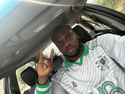 A man is taking a selfie inside a car. The man has dark skin, a beard, and is wearing a white jersey with green accents and the number "10" visible on the left side of the shirt. He is holding up his hand, making a "peace" or "victory" sign. The car has a sunroof, and the interior appears to be black leather. Outside the car window, there are trees and what seems to be a glimpse of a building. Assuming that the location provided is accurate (Lugbe, Nigeria), the image captures a moment of someone inside a car in that area.