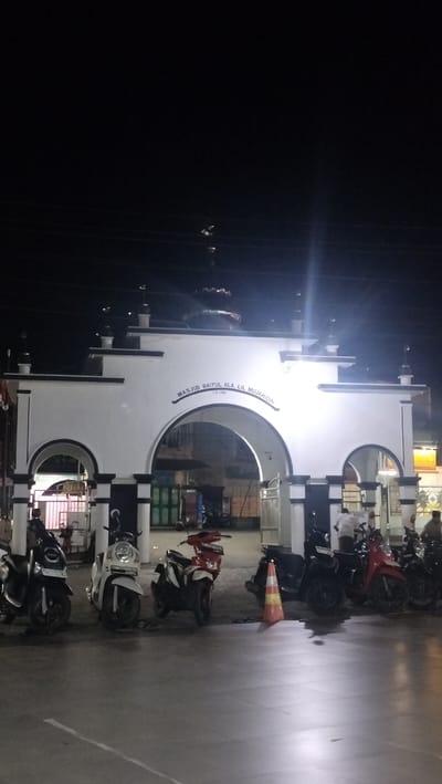 The image shows the entrance to Masjid Baitul Ala Lil Mujahidin in Barat Yaman, Indonesia, at night. The mosque entrance is an arched gateway with a white facade and black accents. Above the archway, the mosque's name is inscribed in Arabic script. Several motorcycles are parked in front of the entrance, illuminated by artificial lighting. Some individuals are visible near the motorcycles and the mosque entrance. The ground is paved, and there are signs of ongoing construction or maintenance in the background. The overall scene is dimly lit, characteristic of a nighttime photograph.