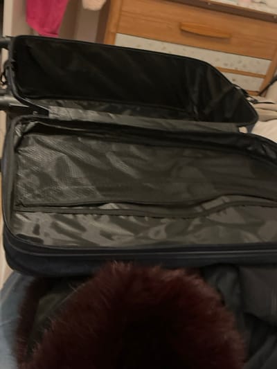 The image displays an open, empty dark-colored soft-sided suitcase, positioned within an indoor environment in the city of Rennes, France. The suitcase's lid is open upwards, revealing a main empty compartment and several internal pockets, some constructed from black mesh material, all lined with a dark grey fabric. In the immediate blurred foreground, a mass of reddish-brown fur-like material is visible at the bottom of the frame. The background features a light-colored wooden dresser or chest of drawers on the right, distinguished by a horizontal handle. Above the dresser, blurred elements including a pink fabric item and a white fluffy object are partially visible. No individuals, explicit actions, or interactions are discernible.