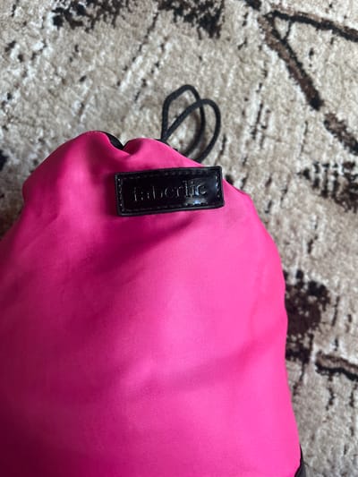 The image shows a close-up, overhead view of a pink drawstring bag resting on a textured carpet. The bag is a bright, solid pink color and appears to be made of a synthetic material, possibly nylon, with a subtle ribbed texture. A black drawstring is visible, coiled loosely to the left of the bag. Attached to the bag is a small, rectangular black tag with a glossy finish. Embossed on the tag in silver lettering are the words "Faberlic". The carpet beneath the bag has a neutral color palette with a pattern of dark brown and grey abstract shapes. The lighting suggests an indoor setting, likely daytime, with no specific environmental cues about the weather or exact location beyond the provided context of Tashlak, Kyrgyzstan. No people are visible in the image. The focus is entirely on the bag and its immediate surroundings.