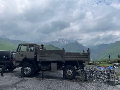 A utilitarian, olive-drab military truck with a flatbed cargo area is parked on a cobblestone surface in Gergeti, Georgia. The truck is positioned in the foreground, with its right side visible. To its left, partially obscured, is a black SUV. The truck has large, rugged tires and appears to be an all-terrain vehicle. In the background, a person stands on a grassy slope overlooking a dramatic mountain landscape with cloudy skies. Piles of stone and blue tarps are visible near the truck. The mountains are steep and green, with patches of snow on their upper reaches. The overall atmosphere is overcast and slightly damp.