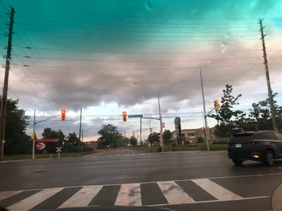 The image depicts an intersection in Barrie, Ontario, Canada, specifically at Big Bay Point Road. The perspective is from inside a vehicle, looking through the windshield, which has some water droplets on it. It is twilight, with overcast skies displaying a gradient from teal to pinkish-grey. Utility poles with numerous wires stretch across the frame.

A traffic light at the intersection shows a red light for the direction of travel. To the left, a large red billboard for "Patene Building Supplies" is visible, along with a railway crossing sign. Several trees line the sides of the road. On the right side of the intersection, a dark-colored SUV is turning, with its right turn signal illuminated. In the background, buildings and more trees are visible, and Canadian flags can be seen mounted on poles. The road surface appears wet, suggesting recent rain.