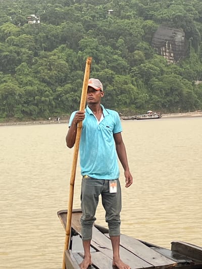 A man stands on a wooden boat in a muddy river, holding a long pole. He is wearing a light blue polo shirt, grey pants, and a baseball cap. The background shows a densely forested hillside with a few buildings and a large rock formation. A boat with passengers is visible on the river in the distance. The weather appears overcast. This scene is typical of the Jaflong area of Bangladesh, known for its natural beauty and riverine landscape.