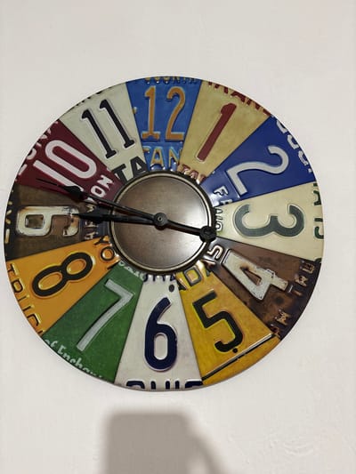A circular wall clock with a distinctive, vintage aesthetic hangs against a plain white wall in an indoor setting in Cuernavaca, Mexico. The clock face is creatively constructed from twelve trapezoidal segments, each designed to resemble a miniature, colorful license plate, displaying the numbers 1 through 12. The metallic central hub of the clock, in a bronze-like finish, anchors three black hands. The minute hand points towards approximately 9, and the hour hand is positioned just past 6, indicating a time of roughly 6:46 or 6:47. A thin black second hand is also visible, pointing between 7 and 8.

Each "license plate" segment features a unique color scheme and number design. For example, the number '2' is set against a blue background with a white number, '3' is on an off-white plate with a dark green number, and '4' is on a rusty brown background with an off-white number. The '7' segment is green with a white number, and partially visible text below it reads "of Enchan", hinting at "Land of Enchantment." Other visible text fragments include "FRA" and "NOA" near the '2', "MTRU" and "MTRN" near the '4', "LA" and "18" near the '5', "NO" near '6' and '10', and "TRUC" and "YOM" near the '8'. The clock casts a soft shadow on the white wall to its right, suggesting ambient indoor lighting.