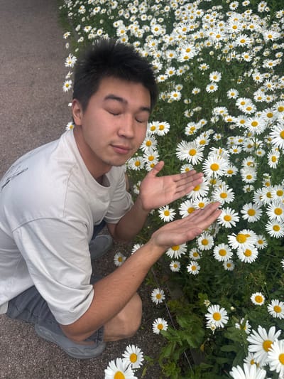 A young East Asian male with dark hair and closed eyes is kneeling in a field of white daisies with yellow centers. He is wearing a light gray t-shirt and dark gray shorts. His hands are extended forward, palms up, as if offering something. The background consists of a dense patch of daisies and green foliage, with a gravel path on the left side. The overall impression is one of peaceful contemplation and appreciation of nature. This scene could be located in the city of Sestroretsk, Russia, which is known for its natural beauty and parks.