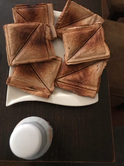 This image presents an overhead view of a static scene on a dark brown wooden table in Calabar, Nigeria. A white, rectangular plate with a curvilinear front edge centrally displays nine triangular halves of toasted sandwiches. The toast coloration ranges from light to medium brown, with localized darker, slightly singed areas visible along the diagonal cuts and perimeters of the crusts. In the lower-left foreground, a grey cylindrical bottle with a white screw-top lid is positioned. Partial orange lettering is discernible on the bottle's side.