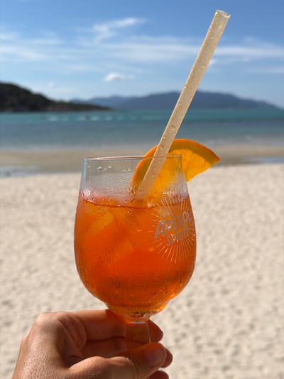 A hand holds a stemmed glass filled with an orange Aperol Spritz. The glass has "APEROL SPRITZ" etched into it and is garnished with an orange slice. A straw extends from the drink, and condensation beads on the outside of the glass. The background is a tropical beach with white sand, turquoise water, and lush green hills under a blue sky with scattered clouds. The location is the beach in Choeng Mon, Thailand.