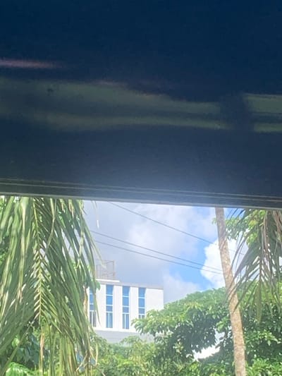 A view from inside a building, possibly through a window or doorway, is dominated by a portion of the structure itself, which is a dark, possibly blue-painted frame. To the right of this frame, the outside world is visible. This includes green foliage from trees and palms, a partly cloudy sky, and a white building with blue-tinted windows. The specific location is Mloganzila, Tanzania. Details about people, actions, or interactions are not provided within the image.