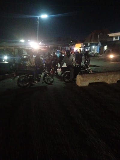 This media file depicts a nighttime scene in Nassarawa, Nigeria. The image is dark and somewhat blurry, suggesting a low-light environment.
The primary subjects are several people, most of whom appear to be on or around motorcycles. In the foreground, two motorcycles are clearly visible, with individuals seated on them or standing nearby. One person is leaning against a motorcycle in the foreground. Further in the background, more people and motorcycles are gathered, creating a sense of activity.
The setting appears to be a street or an open area, possibly a market or a gathering spot, lined with buildings that have lights on. A tall street lamp illuminates a portion of the scene. The presence of motorcycles and people suggests a mode of transportation and social interaction.
The time of day is clearly night, indicated by the darkness and the artificial lighting. There are no visible cues about the weather, but the darkness implies it's not a brightly lit day. The overall atmosphere is one of a bustling, albeit dimly lit, evening gathering. The limited detail due to the image quality prevents the identification of specific emotions or finer details about the clothing or expressions of the people. No visible text is present in the image.