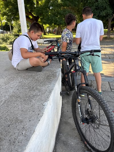 The image depicts three young males outdoors, near bicycles, with trees and a building in the background. The scene appears to be a public park or recreational area.
In the foreground, a boy sits on a concrete ledge, facing left and looking at his smartphone, which he holds in his right hand. He is wearing a white t-shirt, beige shorts, and dark flip-flops. A beige bag is slung over his left shoulder. A black mountain bike with red accents is parked beside him.
To the right of the sitting boy, another boy stands, facing away from the camera and towards the first boy. He is wearing a white t-shirt and light green shorts. His stance suggests he may be interacting with the boy on the phone. Another black mountain bike is positioned in front of him, partially obscuring his legs.
A third boy stands further to the right, also facing away from the camera. He is wearing a white t-shirt with a graphic print and light green shorts. The front wheel of a black mountain bike is prominently visible in the lower right corner of the image.
The environment is characterized by sunlight filtering through dense green trees. The ground consists of a paved area with distinct stone patterns. The concrete ledge the boy is sitting on is weathered. The overall impression is a casual outdoor gathering of young people with their bicycles. The specific location is identified as a city in Kran, Bulgaria.