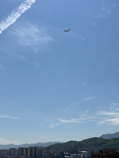 The image shows a plane flying in the sky over Zhangjiajie, China. The plane is in the lower left quadrant of the image and is leaving a contrail. The sky is blue with some wispy clouds. In the right side of the image are buildings and mountains.