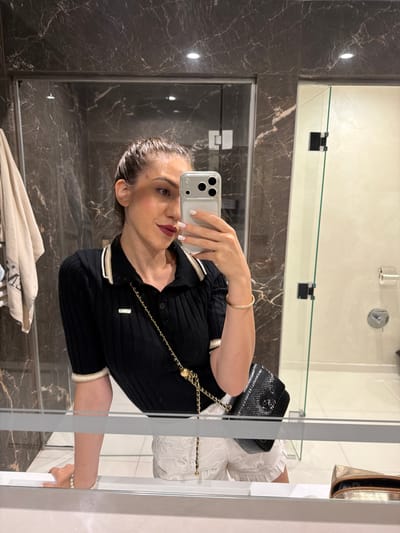 The image is a selfie taken in a bathroom, showing a woman with dark hair pulled back and wearing dark lipstick. She is dressed in a black, short-sleeved, collared top with a ribbed texture and striped trim on the collar and cuffs. A gold chain with a medallion detail is draped across her body, attached to a small black textured handbag. She is also wearing a thin, sparkling bracelet on her wrist.
The background appears to be a modern bathroom with dark marble-patterned walls and a glass shower enclosure. A shelf is visible in the mirror, with a gold cosmetic bag placed on it. The lighting suggests it might be daytime, possibly indoors with artificial lighting. The woman is holding a smartphone to take the photo. There is no visible text in the image.